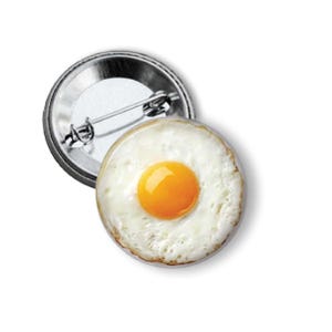 May include: A round button pin featuring a realistic image of a fried egg with a bright orange yolk and white egg whites. The pin has a shiny metal backing with a safety pin clasp.