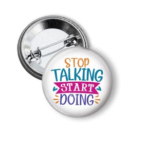 May include: A white button pin with the text "STOP TALKING START DOING" in orange, teal, purple, and pink. The pin has a metal backing with a safety pin closure. The button is round and has a glossy finish.