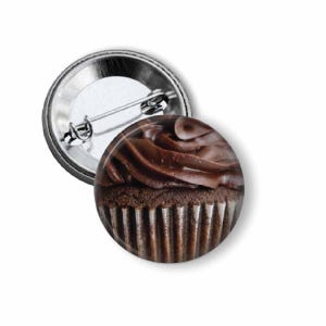 May include: A round button pin featuring a detailed image of a chocolate cupcake with rich, dark frosting. The pin has a silver metal backing with a safety pin closure, perfect for attaching to clothing or bags.