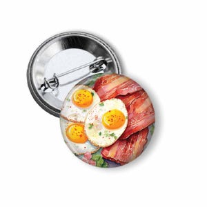 May include: A round button pin featuring a watercolor illustration of a breakfast plate with fried eggs and bacon. The eggs have bright yellow yolks, and the bacon is a reddish-brown color. The pin has a silver metal backing.