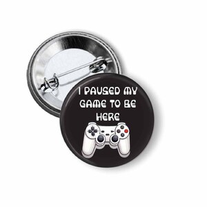 May include: A black button with the text "I paused my game to be here" and a white controller graphic. The button is round and has a metal pin back.