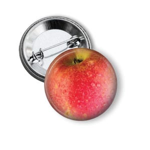 May include: A circular pin-back button featuring a high-resolution image of a red apple with visible water droplets on its skin. The back of the button is made of silver-toned metal and includes a standard pin mechanism for attachment.