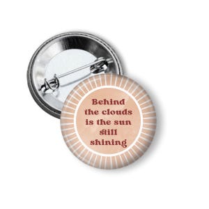 May include: A round button with a pin back. The button face is light brown with a white radial design and the text "Behind the clouds is the sun still shining" in dark brown.