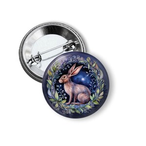 May include: A round button pin with a detailed illustration of a brown hare inside a wreath of green leaves and blue flowers. The background is a dark blue with a starry night sky. The pin has a silver metal backing.