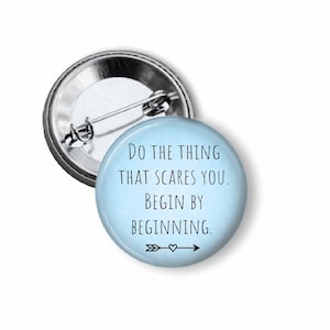 May include: A round button pin with a light blue background and the text "Do the thing that scares you. Begin by beginning." in black. An arrow and heart graphic are at the bottom. The pin has a silver metal backing.