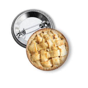 May include: A circular pinback button featuring a high-resolution photographic print of a golden-brown apple pie with a classic lattice crust. The back of the button is made of polished silver-tone metal and includes a standard pin mechanism.