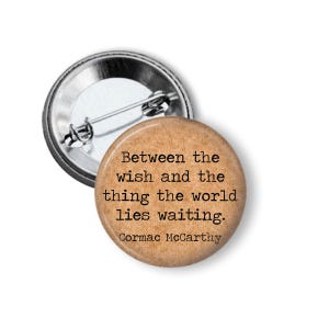 May include: A round button pin with a tan background and the quote "Between the wish and the thing the world lies waiting." by Cormac McCarthy. The pin has a silver metal backing and a safety pin clasp.