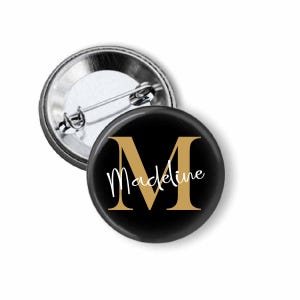 May include: A black button pin with a gold letter "M" and the name "Madeline" in white script. The pin has a silver metal backing with a safety pin closure. The pin is a personalized accessory.