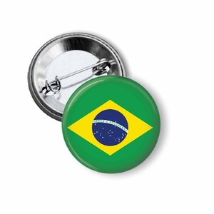 May include: A round button pin featuring the flag of Brazil. The pin has a green background with a yellow diamond in the center. Inside the diamond is a blue circle with white stars and the words "Ordem e Progresso".