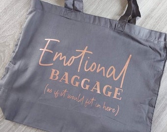 May include: Gray tote bag with the words "Emotional Baggage" in rose gold script. The phrase "(as if it would fit in here)" is below the main text. The bag has two handles and is made of a smooth fabric.