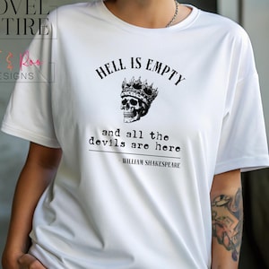 May include: White t-shirt featuring the quote "HELL IS EMPTY and all the devils are here" attributed to William Shakespeare, with a skull wearing a crown graphic. The shirt also has the Novel Attire and JJ & Roo Designs logos.