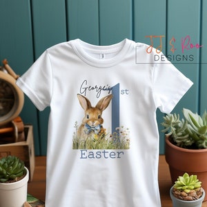 Puede incluir: Camiseta blanca con una ilustraci&oacute;n de un conejito, el n&uacute;mero 1 azul y el texto "George's 1st Easter". El dise&ntilde;o es de estilo acuarela. Ropa infantil con tem&aacute;tica de Pascua.
