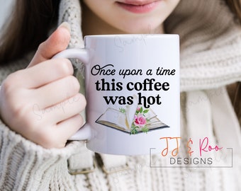 May include: White ceramic mug with a black and white graphic of an open book with pink flowers. The text on the mug reads "Once upon a time this coffee was hot" and "JJ & Roo DESIGNS".