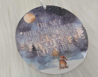 May include: A round Christmas ornament with a winter scene featuring a full moon, snow-covered trees, and a small bird wearing a Santa hat. The text reads, "The sky looks different when someone you love is up there."