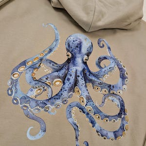 May include: A beige hoodie with a watercolour design of a blue octopus. The octopus has detailed tentacles with light blue and gold accents. The hood is folded down, showing the inside lining.