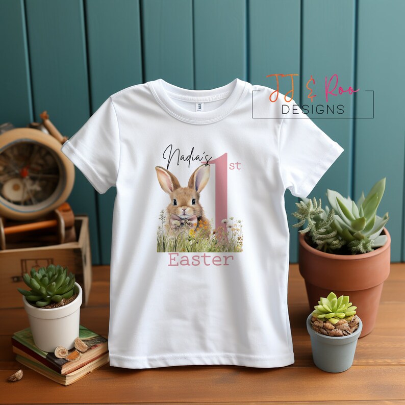 Puede incluir: Camiseta blanca para ni&ntilde;os con un conejo y el texto "Nadia's 1st Easter". El dise&ntilde;o incluye el n&uacute;mero uno en rosa. Una prenda para celebrar la Pascua.