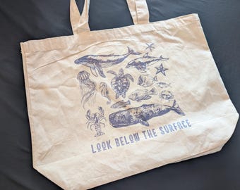 May include: Cream-colored tote bag featuring blue illustrations of marine life, including whales, dolphins, and jellyfish. The text "LOOK BELOW THE SURFACE" is printed at the bottom. The bag has two handles.