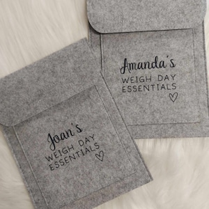 May include: Two gray felt pouches with black text that reads "Joan's WEIGH DAY ESSENTIALS" and "Amanda's WEIGH DAY ESSENTIALS" with a heart symbol. The pouches have a flap closure and a pocket on the front.