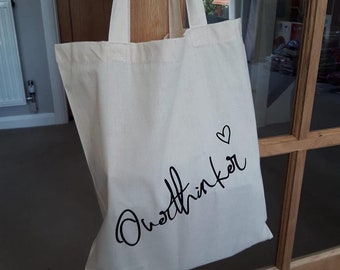 May include: Cream-colored tote bag with black script text that reads "Overthinker" and a small heart symbol. The bag has two handles and is hanging on a wooden door.