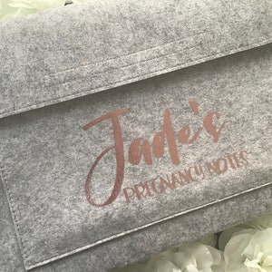 May include: A gray felt folder with a rose gold inscription that reads "Jade's Pregnancy Notes".