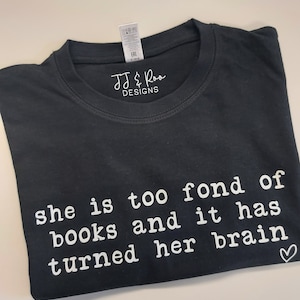 May include: Black t-shirt with white text that reads "she is too fond of books and it has turned her brain" with a small heart symbol at the end.