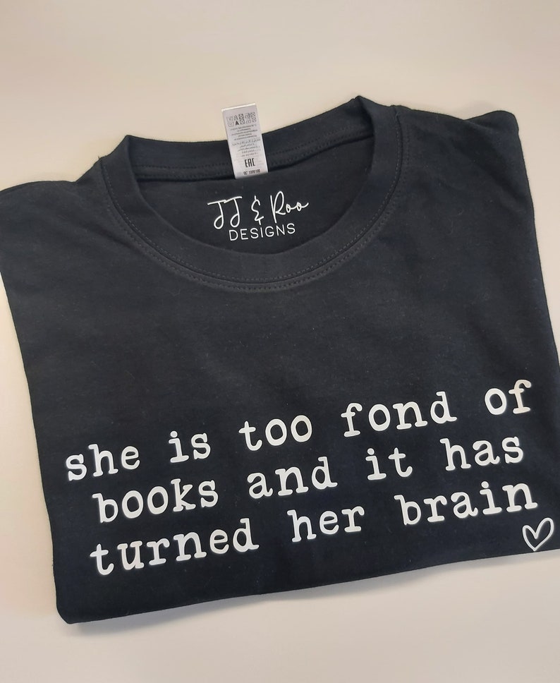 May include: Black t-shirt with white text that reads "she is too fond of books and it has turned her brain" with a small heart symbol at the end.