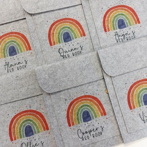 May include: Six grey felt pouches with rainbow designs and the text "[Name]'s Red Book" printed on each. The pouches are stacked on top of each other.