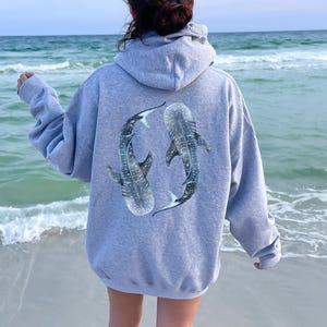 May include: A grey hoodie featuring a graphic of two whale sharks on the back. The hoodie is being worn by a person standing on a beach.