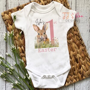 Puede incluir: Body de beb&eacute; blanco con un dise&ntilde;o de conejito y el texto "Isla's 1st Easter". El dise&ntilde;o presenta un conejo marr&oacute;n en un campo de flores, un n&uacute;mero uno rosa y la palabra "Easter" en rosa. El body tiene tres corchetes.
