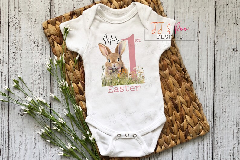 Puede incluir: Body de beb&eacute; blanco con un dise&ntilde;o de conejito y el texto "Isla's 1st Easter". El dise&ntilde;o presenta un conejo marr&oacute;n en un campo de flores, un n&uacute;mero uno rosa y la palabra "Easter" en rosa. El body tiene tres corchetes.