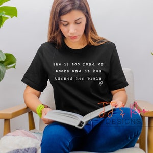 May include: A woman wearing a black t-shirt with the text "she is too fond of books and it has turned her brain &hearts;" printed on it. She is sitting in a chair and reading a book.