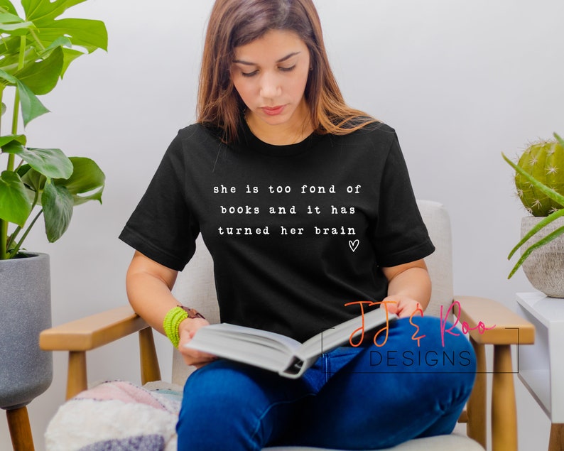 May include: A woman wearing a black t-shirt with the text "she is too fond of books and it has turned her brain &hearts;" printed on it. She is sitting in a chair and reading a book.