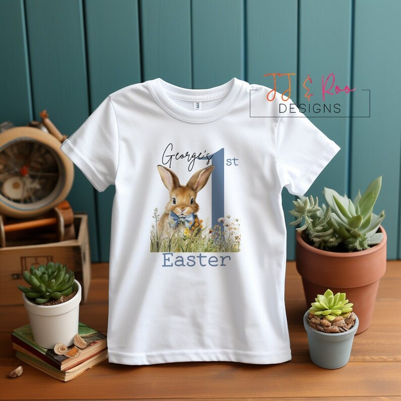 Puede incluir: Camiseta blanca con una ilustraci&oacute;n de un conejito, el n&uacute;mero 1 azul y el texto "George's 1st Easter". El dise&ntilde;o es de estilo acuarela. Ropa infantil con tem&aacute;tica de Pascua.
