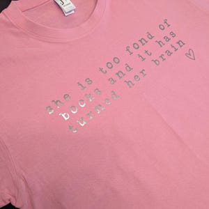 May include: Pink t-shirt with a silver glitter design that says "She is too fond of books and it has turned her brain &hearts;".