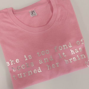May include: Pink t-shirt with silver lettering that reads "She is too fond of books and it has turned her brain"