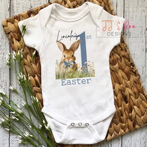 Puede incluir: Body de beb&eacute; blanco con una ilustraci&oacute;n de un conejito, flores y el texto "Lincoln's 1st Easter". El dise&ntilde;o incluye un n&uacute;mero uno azul. El body est&aacute; sobre una estera tejida con flores blancas.