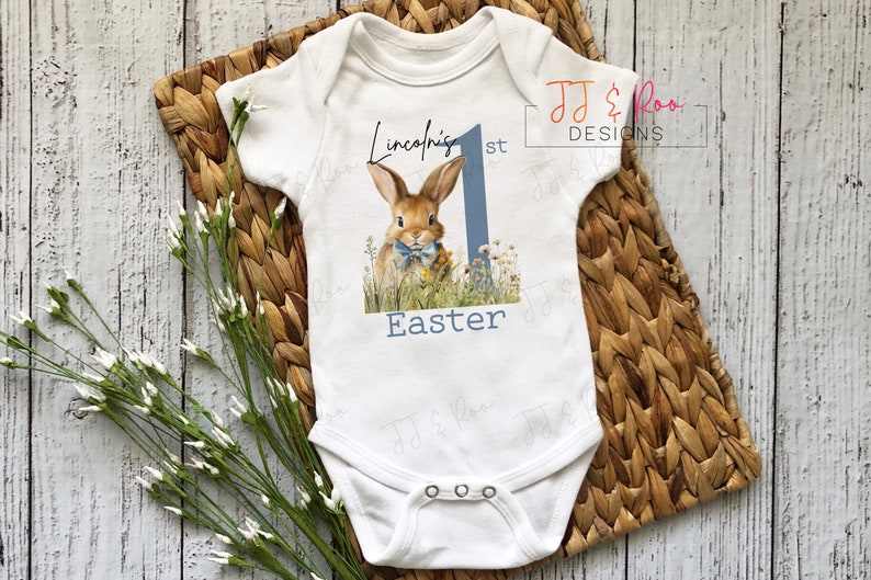Puede incluir: Body de beb&eacute; blanco con una ilustraci&oacute;n de un conejito, flores y el texto "Lincoln's 1st Easter". El dise&ntilde;o incluye un n&uacute;mero uno azul. El body est&aacute; sobre una estera tejida con flores blancas.