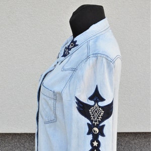 Puede incluir: Una camisa vaquera azul claro con un diseño bordado en blanco y negro en la manga. El diseño presenta un pájaro estilizado con un símbolo yin yang y una estrella.