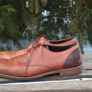 May include: A pair of brown leather lace-up shoes with a brown and orange colour block design. The shoes have a blue trim around the sole.