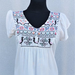 May include: White cotton blouse with short sleeves and a V-neck. The neckline and shoulders feature colorful embroidered designs with birds and geometric patterns.