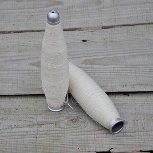 Può includere: Due bobine di filo bianco con tappi metallici su una superficie di legno.