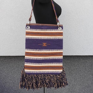 May include: A woven tapestry tote bag with a brown leather strap and fringe trim. The bag has a striped pattern in shades of purple, white, and brown.
