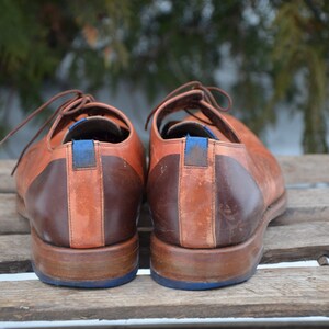 May include: A pair of brown leather shoes with blue accents. The shoes have a two-tone design with a darker brown leather on the heel and a lighter brown leather on the toe.