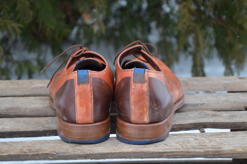 May include: A pair of brown leather shoes with blue accents. The shoes have a two-tone design with a darker brown leather on the heel and a lighter brown leather on the toe.