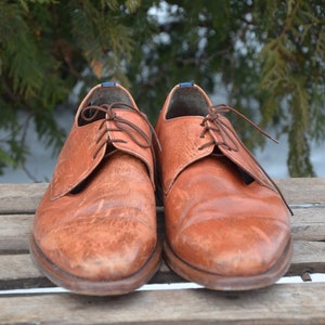May include: A pair of brown leather lace-up shoes with a worn look. The shoes are sitting on a wooden surface.