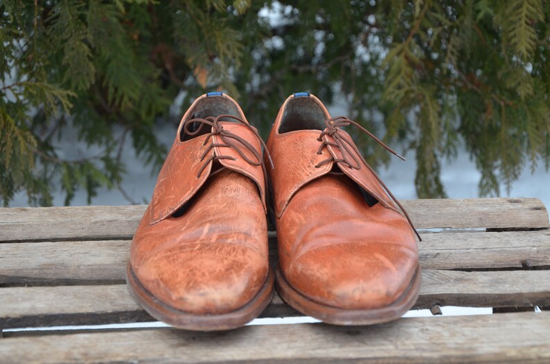 May include: A pair of brown leather lace-up shoes with a worn look. The shoes are sitting on a wooden surface.