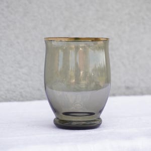 Op de afbeelding: Een transparante glazen tumbler met een gouden rand. Het glas heeft een rokerige grijze tint.