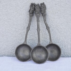Puede incluir: Tres cucharones de plata con mangos adornados con figuras de personas. Los cucharones tienen una forma de cuenco redondeada.