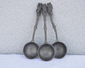 May include: Three silver ladles with ornate handles featuring figures of people. The ladles have a rounded bowl shape.