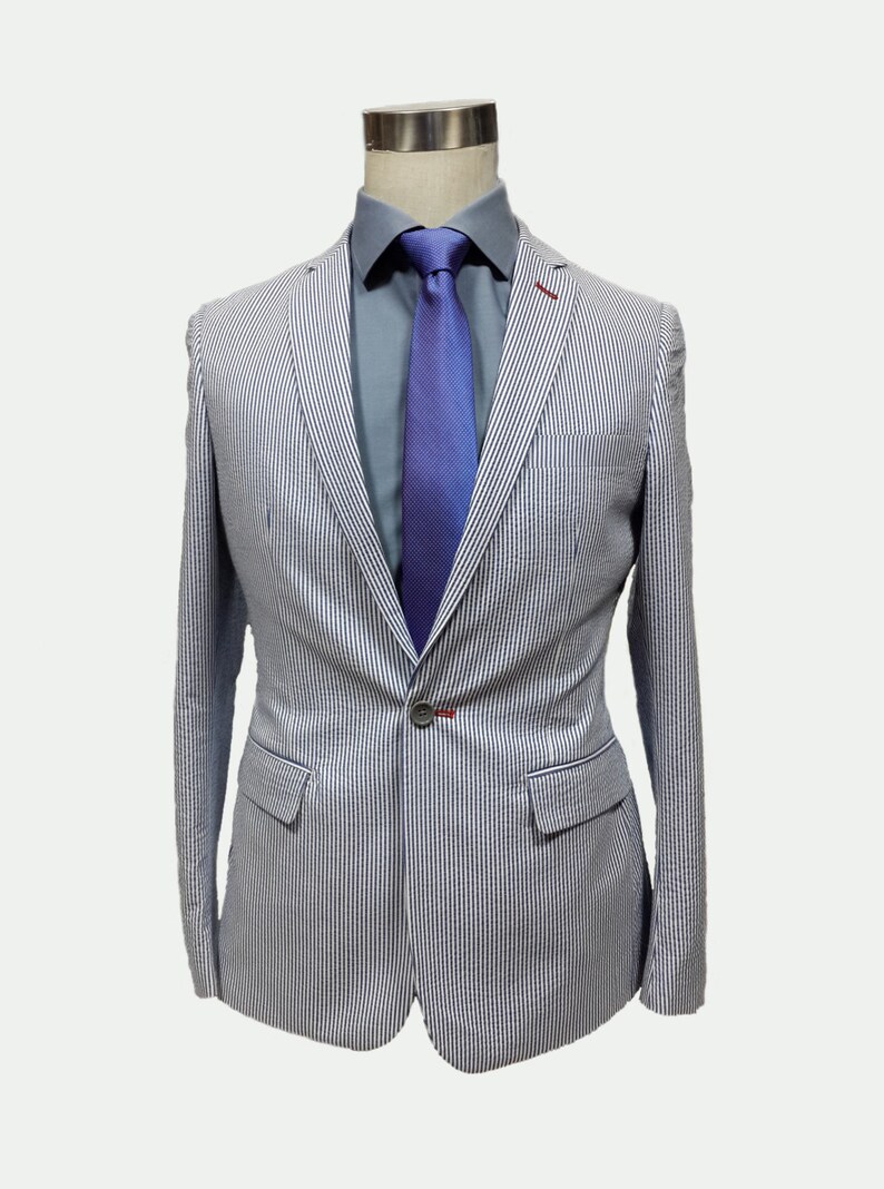 May include: A white and blue striped blazer with a single button closure. The blazer has a light blue shirt underneath with a purple tie.
